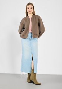 Brown quilted bomber jacket over a light pink top, paired with a light blue denim skirt featuring a front slit, and green suede ankle boots.