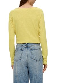 Gelbes Langarmshirt mit lockerem Schnitt und weitem Ausschnitt. Kombiniert mit hochgeschnittenen, hellblauen Jeans mit Gesäßtaschen.