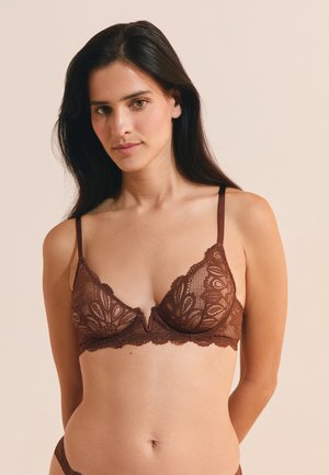 Donna con lunghi capelli scuri che indossa un reggiseno di pizzo marrone, in piedi davanti a uno sfondo beige uniforme.