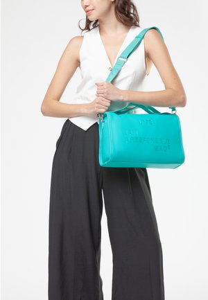 Donna che indossa un gilet bianco e pantaloni neri, con una borsa turchese con la scritta "SONO UNA BORSA RESPONSABILE" sulla spalla.