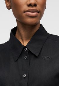 Κοντινό πλάνο ενός ατόμου που φοράει μαύρο πουκάμισο με κουμπιά, αιχμητή γιακά και μικρό κεντημένο λογότυπο στο στήθος.