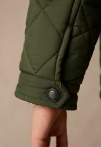 Zaļš dīvāna jaku piedurkne ar redzamu melnu pogu ar zīmolu "SUPERDRY". Gluda virsma, kārtīgas šuves un ģeometriska raksts.