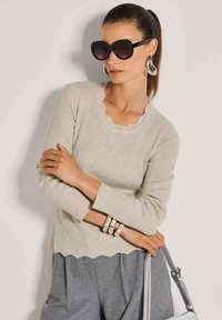Frau trägt große schwarze Sonnenbrillen, silberne Creolen, einen beigen gerippten Pullover, graue Hose, silberne Armbänder und hält eine graue Handtasche.