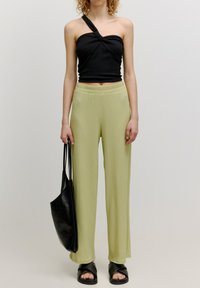 Schwarzes geripptes Top mit einem Shoulder, hellgrüne weit geschnittene Hose mit strukturierter Oberfläche und eine schwarze Ledertasche. Schwarze Rutschsandalen runden das Outfit ab.