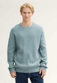 Jasnoniebieski sweter z dzianiny o teksturowanym wzorze, okrągły dekolt oraz ściągacze przy mankietach i u dołu, zestawiony z ciemnoniebieskimi dżinsami.