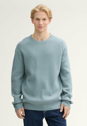 Hellblauer Strickpullover mit einem strukturierten Muster, Rundhalsausschnitt und gerippten Ärmeln sowie Saum, kombiniert mit dunkelblauen Jeans.