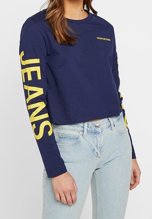 Personne portant un crop top manches longues bleu marine avec un texte jaune "JEANS" sur les manches et un jean taille haute bleu clair.