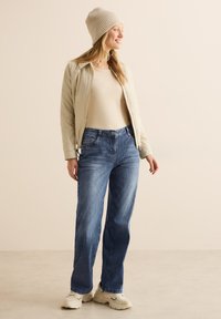 Giubbotto imbottito beige, top beige chiaro, jeans a gamba larga blu e berretto di maglia beige. Le scarpe sono chunky bianche con accenti neri.