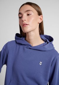Femme à la peau claire et cheveux bruns raides porte un sweat à capuche bleu avec un petit logo blanc en double chevron sur la poitrine.