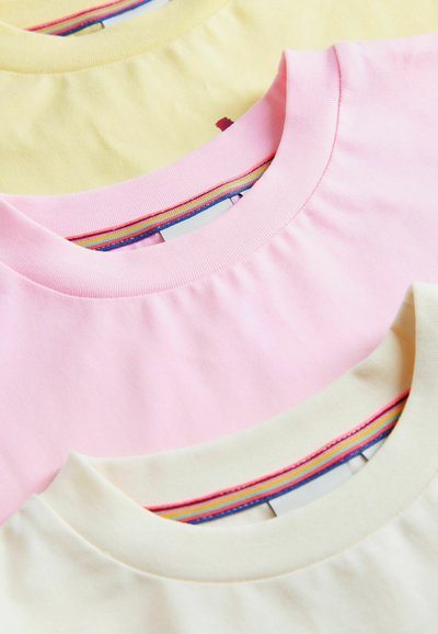Gros plan sur trois t-shirts à col rond pliés en jaune pastel, rose et crème, chacun avec un détail à rayures arc-en-ciel à l’intérieur du col.
