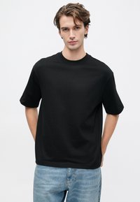 Mladý muž so svetlou pokožkou a hnedými vlasmi, oblečený v jednoduchom čiernom nadmernom tričku a svetlomodrých džínsoch, stojí proti bielej stene.