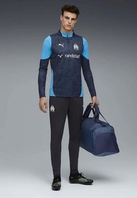 Νέος άνδρας με σκούρο μπλε και ανοιχτό μπλε αθλητικά ρούχα κρατώντας μια μεγάλη ναυτική σακούλα, στέκεται μπροστά σε γκρι φόντο.