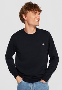 Tengerészkék hosszú ujjú kerek nyakú pulóver puha anyagból, kis logóval a mellrészen, bordázott mandzsettákkal és szegéllyel.