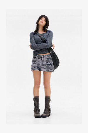 Jovem mulher em pé com os braços cruzados, vestindo uma blusa de manga longa listrada, uma saia mini de camuflagem, botas pretas e uma bolsa preta contra um fundo branco.