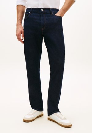 Homme portant un jean droit bleu foncé avec une chemise blanche boutonnée et des baskets blanches, une main dans la poche.