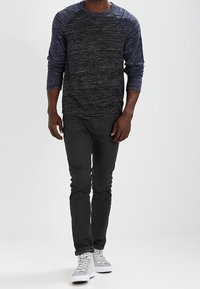 Chemise à manches longues gris foncé avec des manches raglan bleues, associée à un pantalon slim noir et des sneakers montantes blanches. Tissu doux et texturé.