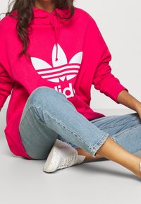 Osoba v jasně růžové mikině Adidas, světlých modrých džínách a bílých teniskách, sedící v přehnuté pozici proti jednobarevnému pozadí.