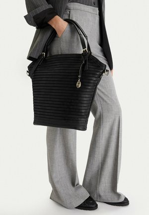 Persona vistiendo blazer y pantalones gris con rayas finas, sosteniendo un bolso grande negro con textura, con asas dobles y un colgante dorado.