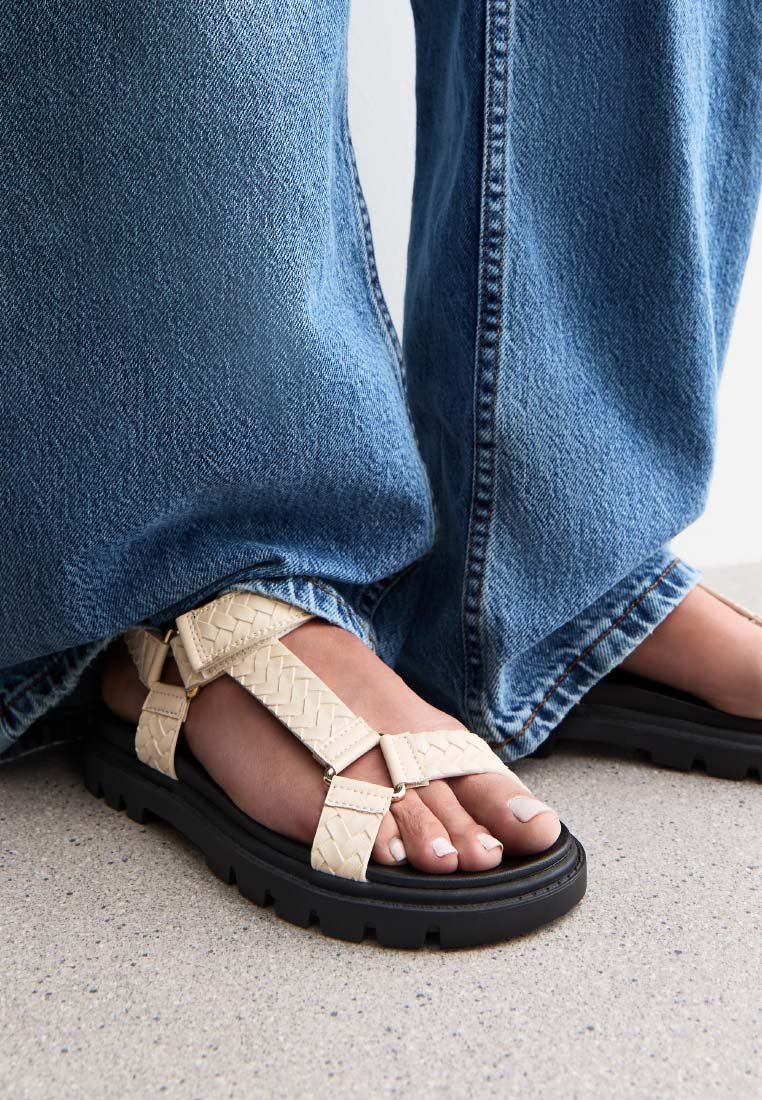 Sandali intrecciati beige con cinturini larghi, suola in gomma nera e accenti testurizzati. Abbinati a jeans di denim blu.
