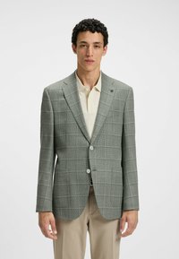 Grå rutiga kavaj i texturerat tyg, med två knappar, slagkrage och sidofickor, bärs över en ljusbeige polo-tröja.