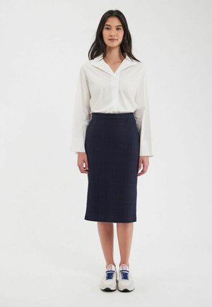 Blouse blanche à col montant avec des manches longues, associée à une jupe texturée bleu marine longueur genou et des baskets blanches avec des accents bleus.