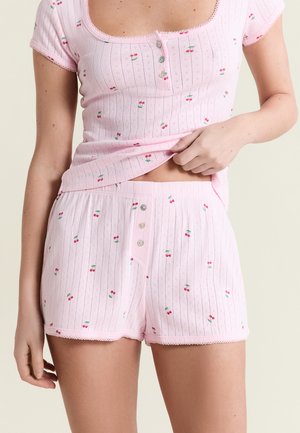 Kobieta w różowych prążkowanych szortach i dopasowanym topie z krótkim rękawem z nadrukiem wiśni, lekko podnosząca top, aby pokazać okolicę talii.