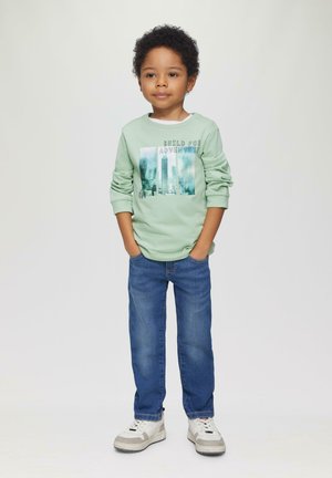 Lichtgroen longsleeve shirt met de tekst "Bouw voor Avontuur", gecombineerd met blauwe jeans en witte sneakers. Handen in de zakken.