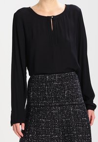 Schwarze Bluse mit langen Ärmeln und runder Ausschnittöffnung, die ein Schlüsselloch hat und mit dekorativen Nähten versehen ist. Kombiniert mit einem strukturierten dunklen Rock.