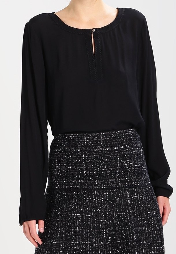 Schwarze Bluse mit langen Ärmeln und runder Ausschnittöffnung, die ein Schlüsselloch hat und mit dekorativen Nähten versehen ist. Kombiniert mit einem strukturierten dunklen Rock.