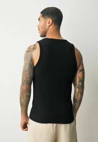 Sort, ribbet tanktop med rund hals og brede skuldre, båret af en person med synlige tatoveringer, parret med lyse shorts.
