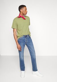 Groene katoenen t-shirt met korte mouwen, blauwe denim jeans en witte sneakers. Een rode bandana om de nek voegt een vleugje kleur toe.