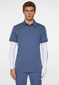 Blå pikétröja med standardkrage, korta ärmar och en logotyp på kragen; bärs över en vit långärmad skjorta och grå handskar.