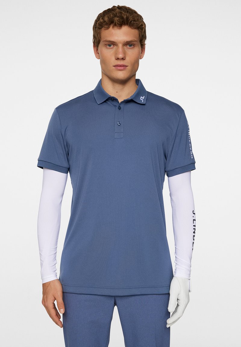 Blå pikétröja med standardkrage, korta ärmar och en logotyp på kragen; bärs över en vit långärmad skjorta och grå handskar.