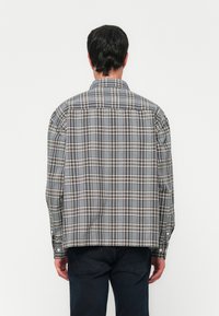 Man gezien vanaf de achterkant, gekleed in een grijs geruite lange mouwen shirt en donkere broek, tegen een effen witte achtergrond.