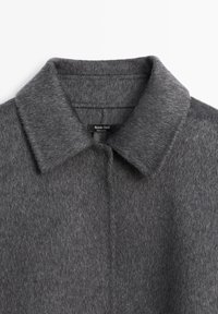 Manteau gris en mélange de laine avec col à revers, texture douce et design simple avec fermeture centrale à boutons. Étiquette de la marque visible.