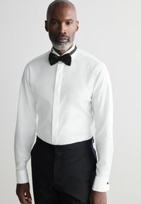 Camicia bianca con un papillon nero a contrasto. Presenta un davanti con bottoni e polsini doppi. Indossata con pantaloni neri su misura.