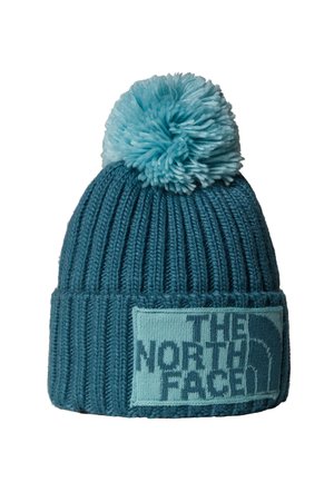 Gebreide muts in teal, met een grote pompon bovenop, geribbelde textuur en een rechthoekige patch met het "THE NORTH FACE" logo in lichtblauw.