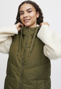 Olivový puffer vest s vysokým golierom, predným zipsom a šnúrkou. Nosený cez textúrovaný krémový sveter so širokými rukávmi.