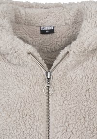 Beige pehmeä fleecetakki hopeisella vetoketjulla ja pyöreällä vetimellä, jossa sisäkauluksessa koko XS ja teksti "CLASSICS".