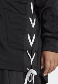 Musta takki, jossa valkoiset nyörit sivuilla, hopeiset silmukat ja teksturoitu kangas. Tarkoituksena on korostaa leveää hihansuun ja taskun yksityiskohtaa.