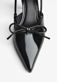 Čierna lodička z lakovanej kože s lesklým povrchom, má úzky remienok a ozdobnú mašľu vpredu.