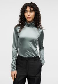 Μπλούζα με μακριά μανίκια και ζιβάγκο σε απαλό τιρκουάζ σατέν με λείο ύφασμα, συνδυασμένη με μαύρο, κομψό παντελόνι με στενή εφαρμογή και διακριτική λάμψη.