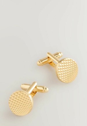 Une paire de boutons de manchette ronds en or avec un motif de pyramide texturé sur les faces, reposant sur une surface gris clair.