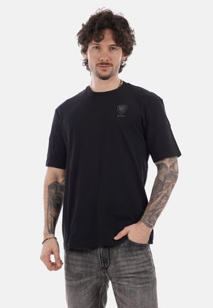 Giovane uomo con capelli ricci, che indossa una t-shirt nera NYPD e jeans grigi, con maniche tatuate complete su entrambe le braccia, in piedi con una mano in tasca.