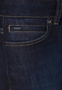 Vaqueros de denim azul oscuro con costuras visibles, un pequeño bolsillo frontal y un botón de metal. La textura parece suave con un ligero brillo.