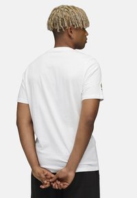 Hvid bomuld t-shirt med korte ærmer, standard rund hals, ensfarvet ryg og subtilt logo detaljeret på venstre ærme. Glat tekstur.