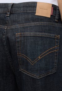 Σκούρο μπλε τζιν από denim με πορτοκαλί ραφές και δερμάτινο ταμπελάκι με την επιγραφή MOS MOSH στη μέση, φορεμένα με λευκό τοπ.