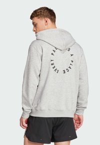 Grå huvtröja med en cirkulär svart textdesign på baksidan. Har en avslappnad passform och ribbade muddar. Svarta shorts syns nedanför.