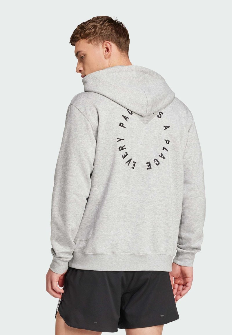 Grå huvtröja med en cirkulär svart textdesign på baksidan. Har en avslappnad passform och ribbade muddar. Svarta shorts syns nedanför.