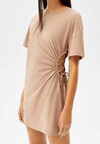 Beżowa sukienka z krótkim rękawem, z marszczeniami po bokach i ozdobą w postaci sznurowania. Wykonana z miękkiego materiału, zapewnia luźne dopasowanie i casualowy styl.